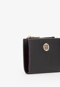 Portafoglio in pelle nera con zip dorata ed emblema circolare dorato sulla parte anteriore, visibile in vista laterale con foderatura interna rosa.