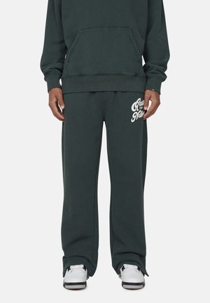 Felpa verde scuro con tasca frontale, abbinata a pantaloni della tuta coordinati con testo grafico bianco. Vestibilità comoda e rilassata.