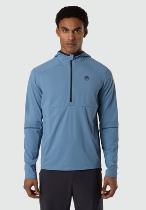 Uomo che indossa una felpa blu a maniche lunghe con mezza cerniera e pantaloni scuri, in piedi rivolto verso l'obiettivo su uno sfondo chiaro e uniforme.