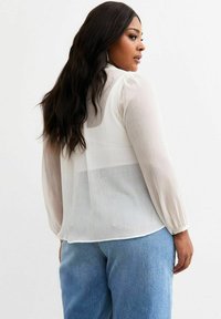 Weißes, durchscheinendes Langarmblusenshirt mit plissierter Textur, runder Ausschnitt und lockerer Passform, kombiniert mit hellblauen Jeans.
