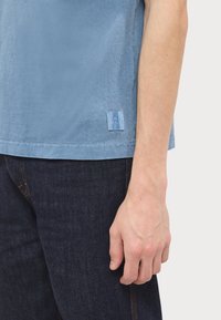 Bras droit et main d'une personne portant une chemise à manches courtes bleue et un jean bleu foncé sur un fond clair uni.