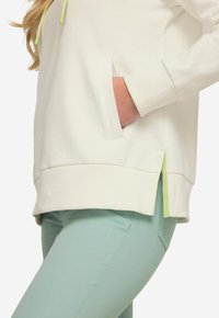 Felpa bianca con polsini a costine e uno spacco laterale che presenta un accento verde chiaro a contrasto. Abbinata a pantaloni verde chiaro.