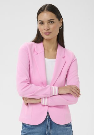 Kvinde med langt mørkt hår iført guldringe i ørerne, en lyserød blazer med stribede manchetter, hvid bluse og blå jeans, armene krydsede.