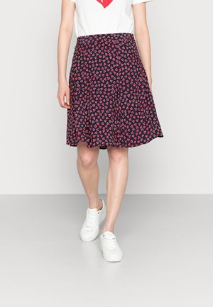 Jupe marine avec une forme évasée, ornée d'un motif répétitif de petits cœurs rouges, confectionnée dans un tissu léger. Portée avec des baskets blanches.