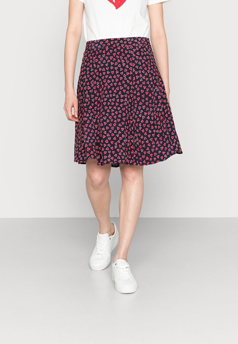 Gonna blu con una forma svasata, caratterizzata da un motivo ripetuto di piccoli cuori rossi, realizzata in tessuto leggero. Indossata con sneakers bianche.