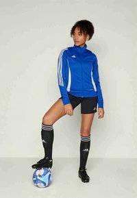 Γυναίκα με μπλε αθλητικό μπουφάν, μαύρες βερμούδες και κάλτσες, να στέκεται με το ένα πόδι πάνω σε μια μπλε και άσπρη μπάλα ποδοσφαίρου μπροστά από έναν απλό φόντο.