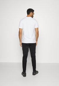 Weißes, kurzärmliges T-Shirt und schwarze Slim-Fit-Hosen. Der Stoff wirkt glatt; das T-Shirt hat einen runden Ausschnitt, die Hose hat eine Reißverschlusstasche.