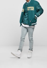 Tealowa kurtka varsity z żółtym napisem, warstwowo nałożona na tealowy sweatshirt, w połączeniu z jasnoniebieskimi skinny jeansami i białymi sneakersami na wysokiej podeszwie.