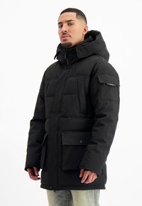 Man in een zwarte gewatteerde winterjas met grote voor- en mouwzakken, staand tegen een effen witte achtergrond.