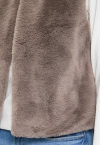 Weicher brauner Kunstfell-Westen, getragen über einem weißen gerippten Pullover und blauen Jeans, die Textur und Schichtdetails zeigen.