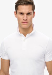 Biała koszulka polo z krótkim rękawem i zapięciem henley na guziki. Materiał wydaje się gładki i dopasowany, z małym logo na lewym rękawie.