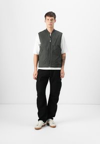 Chaleco sin mangas de color verde oliva con cremallera frontal, dos bolsillos laterales, llevado sobre una camiseta blanca, combinado con pantalones cargo negros y zapatillas blancas.