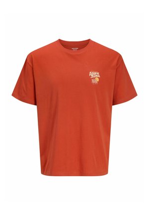 Dreng iført en orange t-shirt med tekst, lyseblå lange cowboyshorts, hvide sokker og beige sko, der står og smiler.