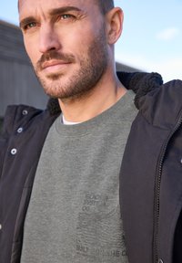 Grå sweatshirt med präglade grafik, texturerat tyg och rundad hals, under en svart zippered huva jacka med fleecefoder.