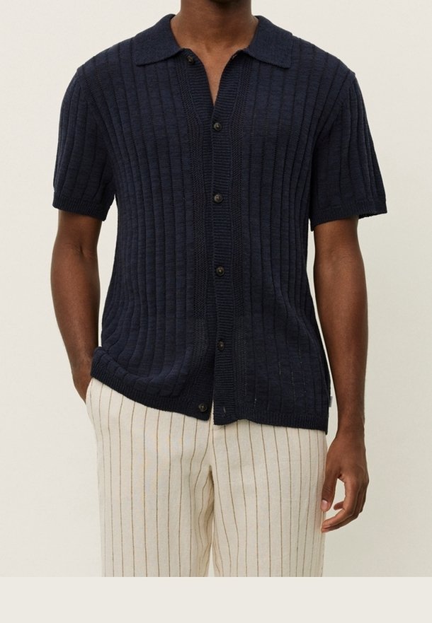 Homme portant une chemise tricotée à manches courtes bleu marine boutonnée et un pantalon crème avec de fines rayures verticales marron.