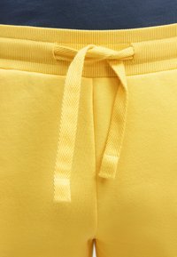 Gelbe Jogginghose mit einem gerippten Bund und gebundenem Kordelzug, getragen über einem dunkelblauen Oberteil.