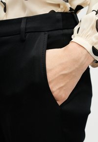 Main dans la poche d'un pantalon noir sur mesure, associée à un chemisier crème à pois noirs avec poignet froncé.