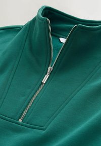 Grøn fleece-pullover med høj krave, der har en sølvfarvet lynlås og en subtil tekstureret overflade. Inkluderer en frontlomme med flad søm.