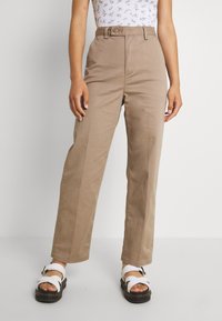 Ljusbruna högmidjade byxor med veck framtill, sidofickor och rak bendesign. Matchas med vita sandaler.