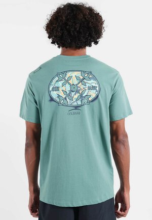Homme aux cheveux bouclés portant un t-shirt turquoise avec un motif floral comprenant des flèches croisées et le texte « Restez en vie » et « Oxbow » au dos.