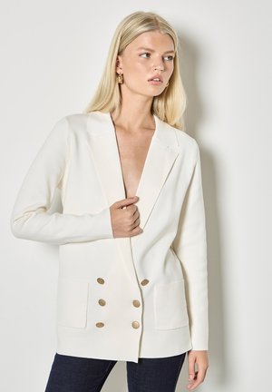 Mujer rubia con una blazer blanca de doble botonadura y botones dorados, y pantalones oscuros, de pie frente a un fondo gris claro.