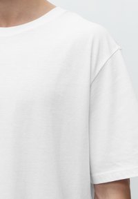 Person wearing a plain white short-sleeve crew neck t-shirt, visible from neck to mid-arm against a light background.