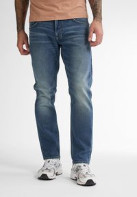 Jeans azzurri chiari con taglio dritto, caratterizzati da un leggero scolorimento, design a cinque tasche e cuciture a contrasto, indossati con scarpe da ginnastica bianche.