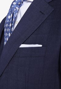 Granatowa marynarka w delikatną kratę, biała koszula, niebieski krawat w wzory oraz biały poszetka w kieszeni piersiowej.