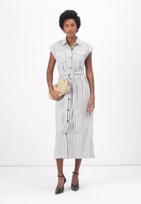 Rochie cu dungi, neagră și albă, cu talie accesorizată cu o curea, tivul până la genunchi și mâneci scurte, plisate. Femeie care ține o poșetă.