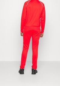 Tuta rossa con design aderente, caratterizzata da strisce laterali bianche e polsini a costine. Il materiale sembra liscio ed elastico, abbinato a scarpe da ginnastica nere.