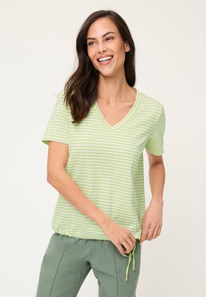 Donna sorridente che indossa una maglietta a righe verde chiaro e bianca con scollo a V, orlo con coulisse e pantaloni verdi larghi, in piedi davanti a uno sfondo neutro.