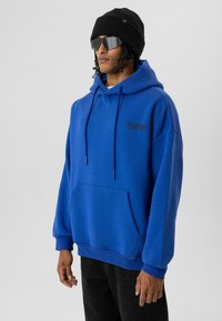 Blauwe pullover hoodie van zacht materiaal, met een voorvak in kangoeroe-stijl, trekkoorden en een zwart geborduurd logo op de borst.