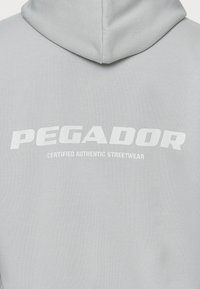 Felpa grigia con cappuccio caratterizzata da una grande stampa bianca che riporta la scritta "PEGADOR" con il sottotitolo "CERTIFIED AUTHENTIC STREETWEAR" sotto. Texture liscia.