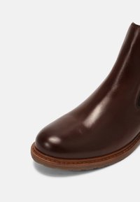 Stivale alla caviglia in pelle marrone con finitura lucida, punta tondeggiante, pannelli laterali elasticizzati e suola in gomma beige. Texture liscia e dettagli minimi.