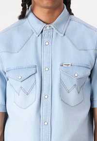 Lys blå denimskjorte med korte ermer, med to frontlommer med spisse klapper, hvite sømdetaljer og trykknapper.
