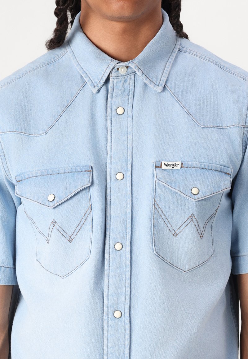 Jasnoniebieska koszula dżinsowa z krótkim rękawem, z dwoma przednimi kieszeniami z odstającymi klapami, białymi detalami szwów oraz zapięciem na napy.