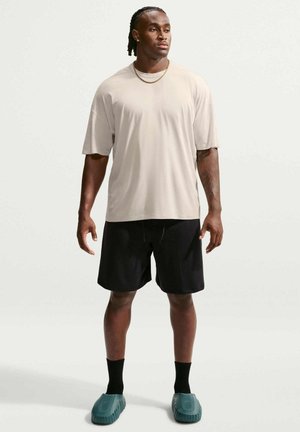 Uomo che indossa una t-shirt beige oversize, pantaloncini neri, calzini neri, scarpe slip-on turchesi e una catena d'oro, in piedi davanti a uno sfondo chiaro e uniforme.