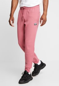 Pink sweatpants made of soft fabric, featuring a drawstring waist, side pockets, and a small black logo patch on the thigh.
