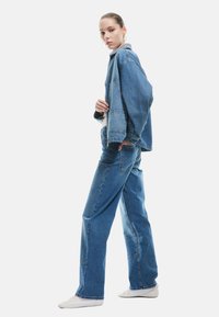 Giacca di jeans azzurro chiaro e jeans a vita alta, vestibilità rilassata, con cuciture visibili e orlo leggermente sfilacciato. Indossata con sneakers bianche.