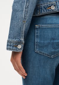 Κλείσιμο του χεριού ενός ατόμου κοντά στο μανίκι ενός μπλε τζιν μπουφάν με ασημένιο κουμπί, δίπλα στην πίσω τσέπη μπλε τζιν παντελονιού.