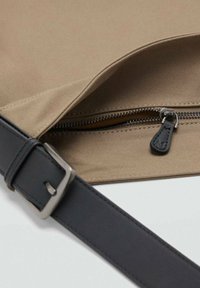 Bolsa de tela color marrón claro con una correa de cuero negra. Cuenta con un cierre de cremallera con un tirador plateado y costuras finas. El interior está forrado en amarillo.