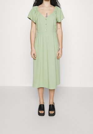Robe midi en tissu vert clair, avec des manches bouffantes, un décolleté en V profond, des boutons sur le devant et une jupe fluide et droite. Sandales à plateforme noires.