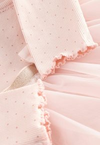 Jemně růžová látka s malým tečkovaným vzorem, vroubkovaná textura, rýhující okraje a vrstvený průsvitný síťový detail se subtilním metalickým pruhem.