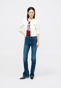 Cardigan branco e texturizado com botões dourados, combinado com uma t-shirt gráfica em bordô e jeans flare azul-escuro. Modelo está de pé com as mãos na cintura.