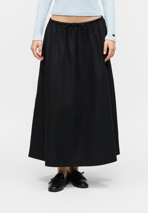 Naine, kellel on must nöörvööga A-lõikega maksiseelik ja mustad paeltega kingad ning helesinine pikakarvaline pluus, seisab valge tausta ees.