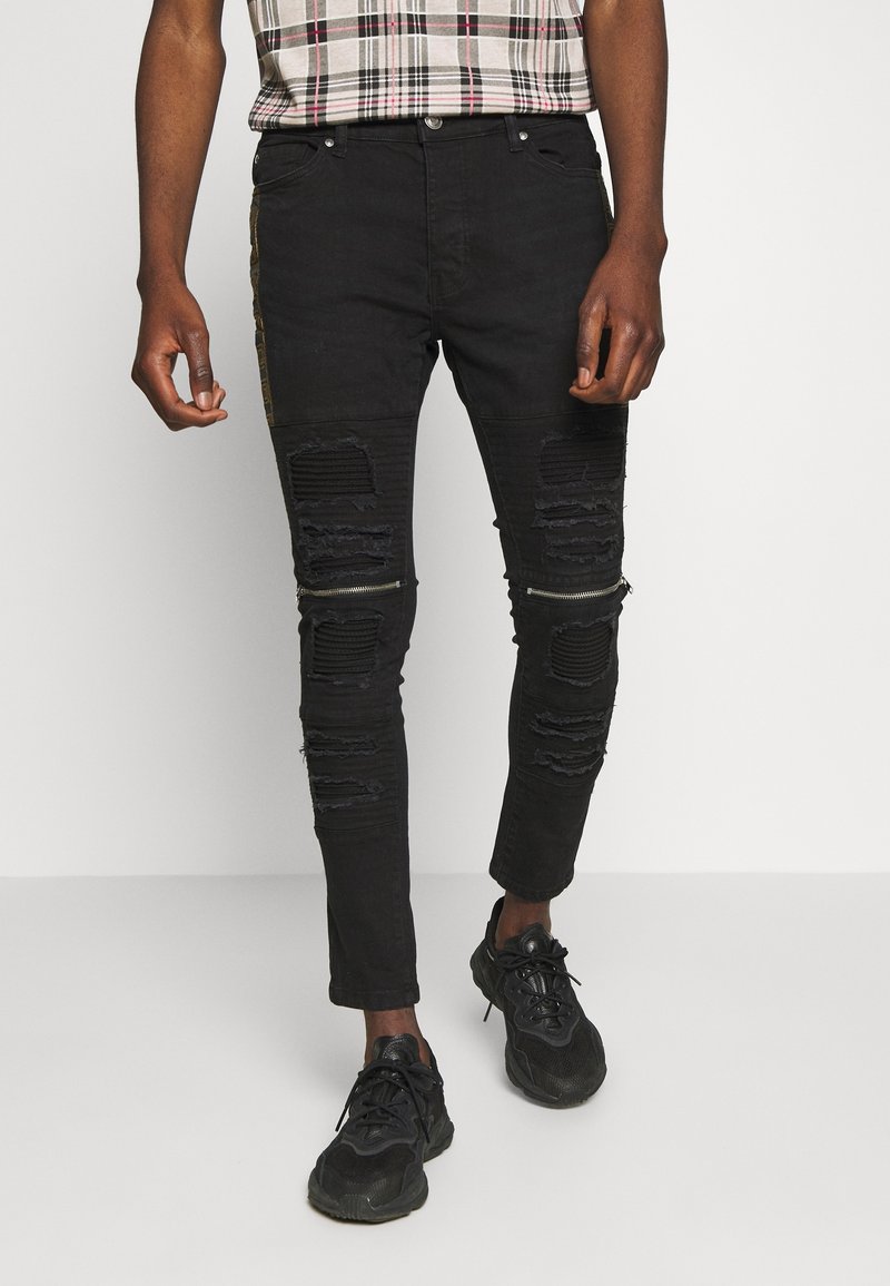 Vaqueros negros ajustados con detalles desgastados, que presentan desgarros en las rodillas, un diseño texturizado y cremalleras metálicas en los lados.