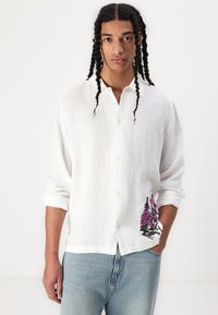 White linen shirt with a relaxed fit, featuring a floral embroidery in purple on the left pocket, and rolled-up sleeves.