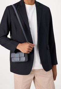 Graue Umhängetasche mit rechteckiger Form und strukturierter Oberfläche, ausgestattet mit einer vorderen Klappe und einem minimalistischen Schnallendetail, getragen über einem schwarzen Nadelstreifenblazer.