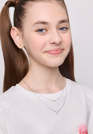 Dwie srebrne naszyjniki nałożone na białą koszulę. Jeden z nich ma zawieszkę w kształcie motyla, a drugi prosty geometryczny kształt. Brązowe włosy, jasna karnacja.