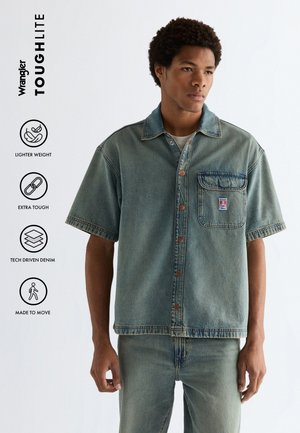 Mann iført kortermet denimskjorte med knapper og jeans, stående mot en ensfarget bakgrunn med fire ikoner som fremhever stoffets egenskaper.